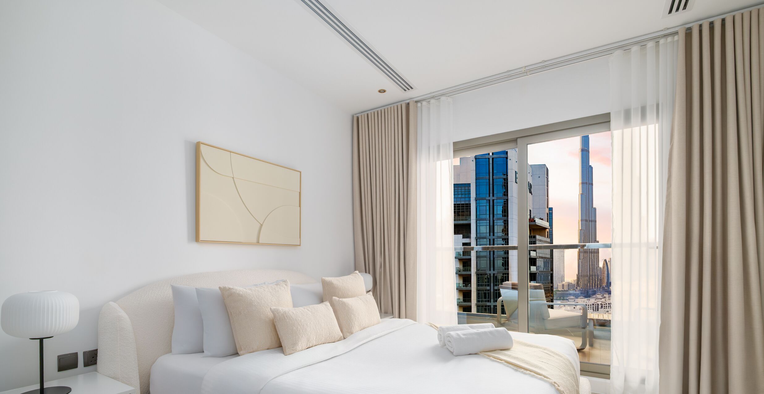 A modern hotel room with a large bed, a bedside table, a lamp, a city view through a large window, and a piece of abstract art on the wall.