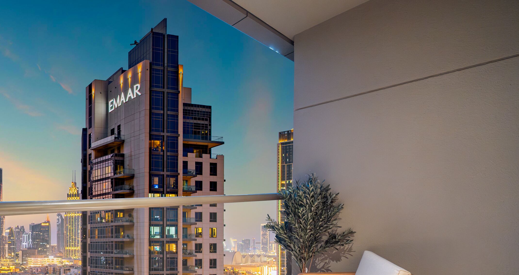 A modern building with the name "EMMAAR" on its facade, viewed from inside a room with a balcony overlooking a cityscape at dusk.