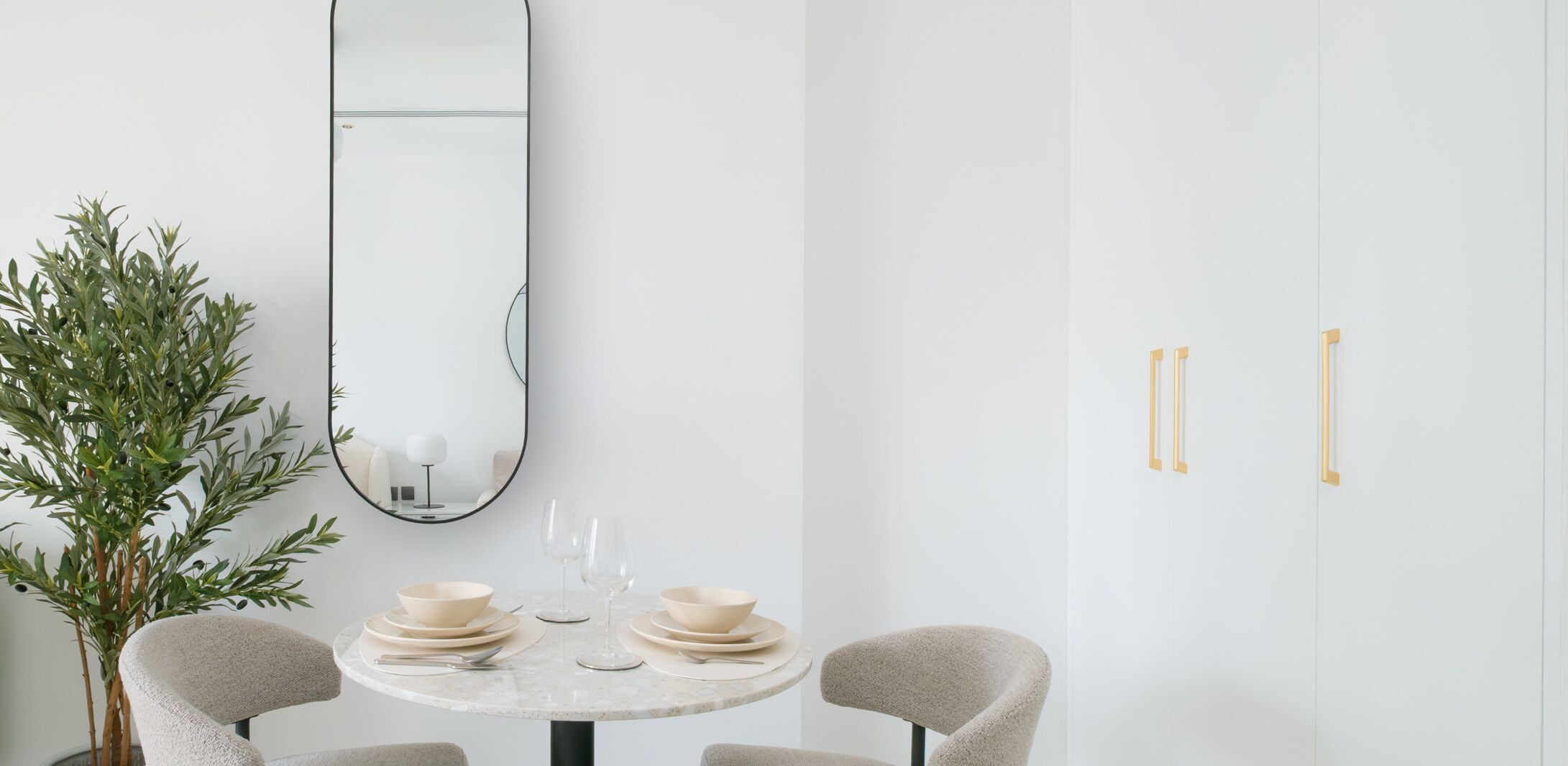 Un coin repas minimaliste avec une table ronde, deux chaises, un miroir et une plante en pot.
