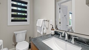 A bathroom with a toilet, a sink, a mirror, and a window with blinds. - Charming 2-bedroom studio with WiFi, AC in the heart of downtown Bartlesville (Bartlesville)
