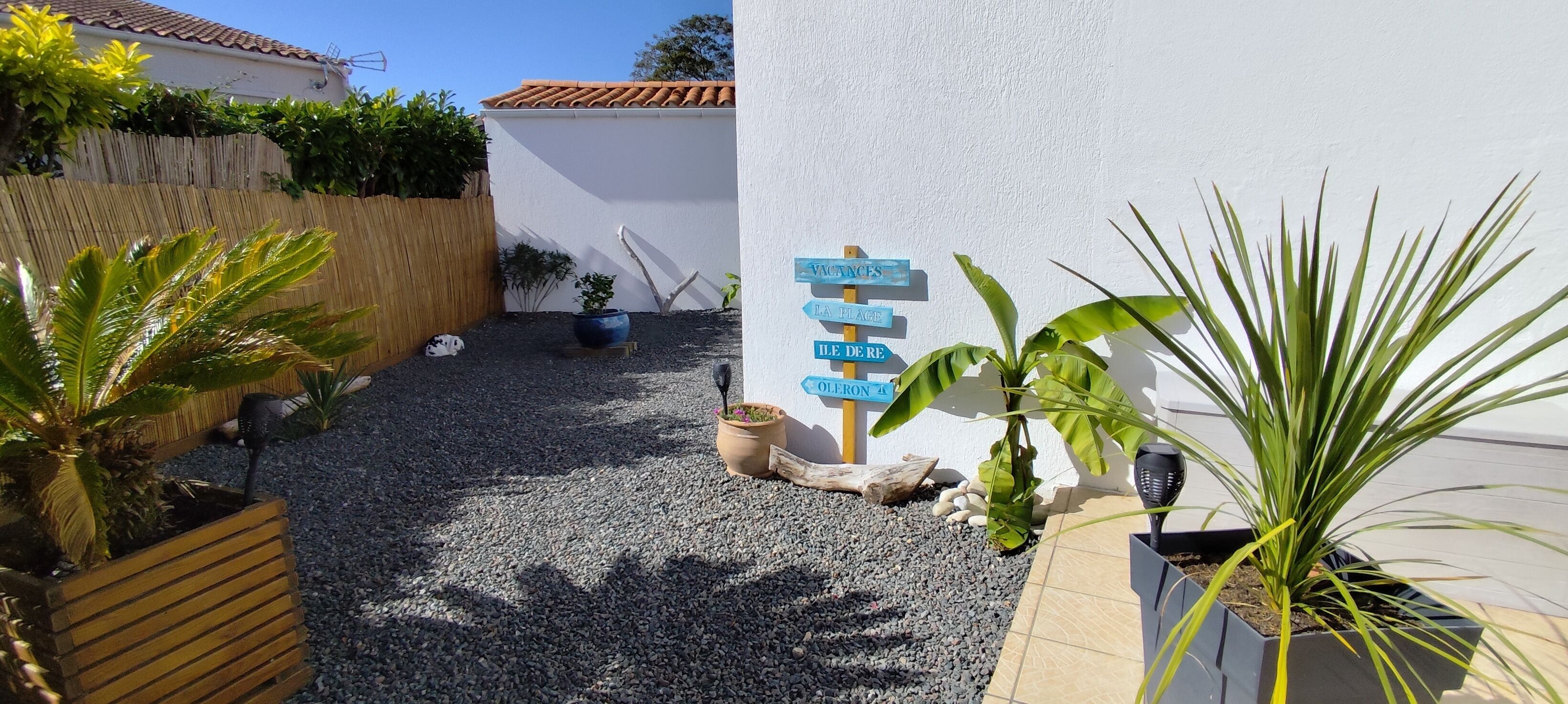 Un jardin avec un chemin de gravier, une clôture en bois, des plantes en pot et des panneaux indicateurs.