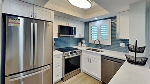 Una cocina moderna con electrodomésticos de acero inoxidable, armarios blancos, un salpicadero de azulejos azules y una cesta colgante negra sobre la encimera.
