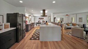 Una cocina moderna con una isla central, un comedor con una mesa larga y una sala de estar con un sofá y un televisor.