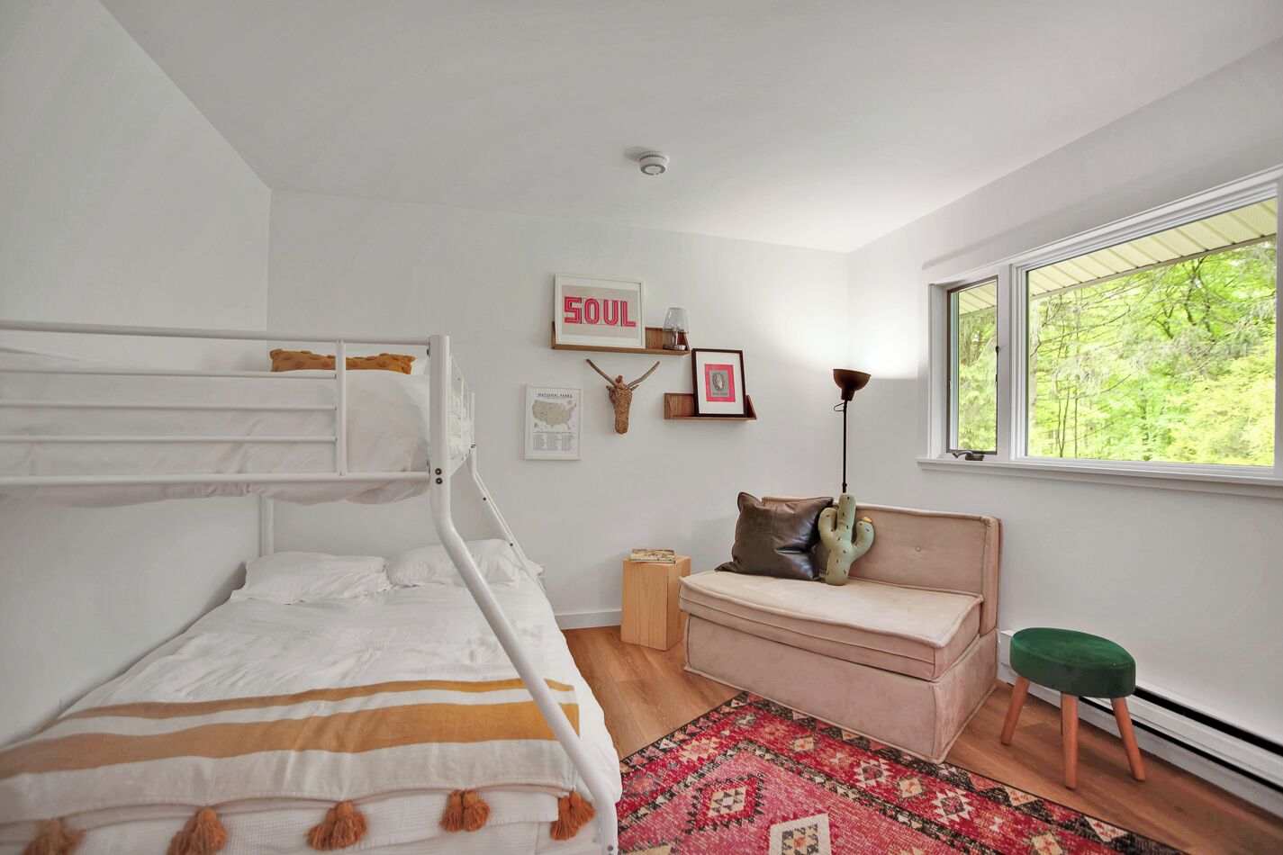A bedroom with a bunk bed, a sofa, a cactus decoration, a rug, and a window with a view of trees.