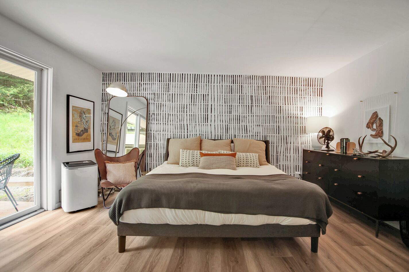A modern bedroom with a large bed, a chair, a dresser, and a window with a view of greenery.
