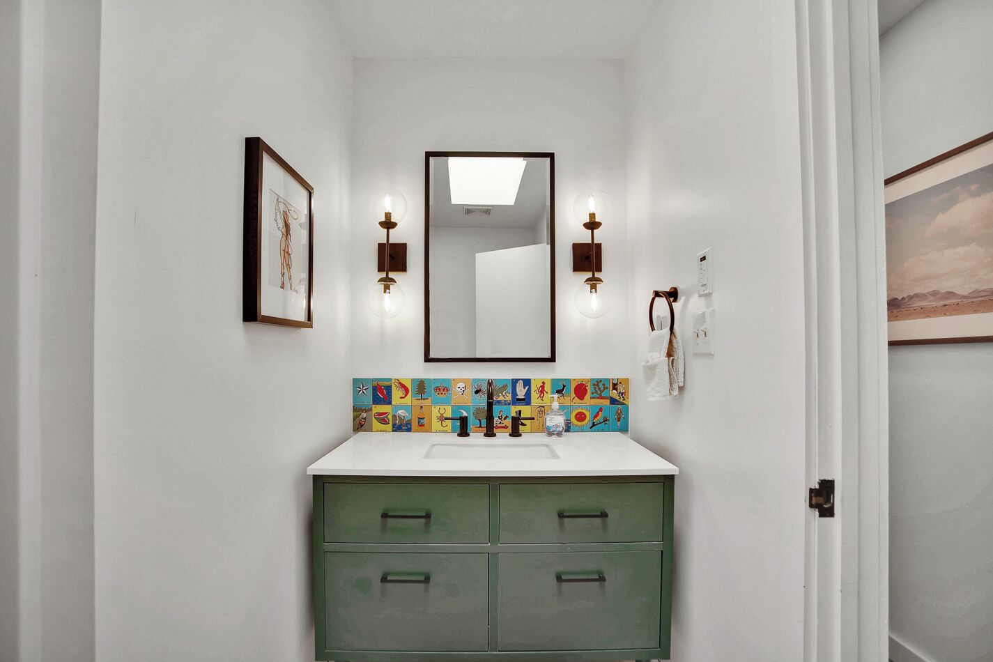 Une salle de bain avec un mur blanc, un meuble vert, un pare-feu coloré, un miroir et deux appliques murales.