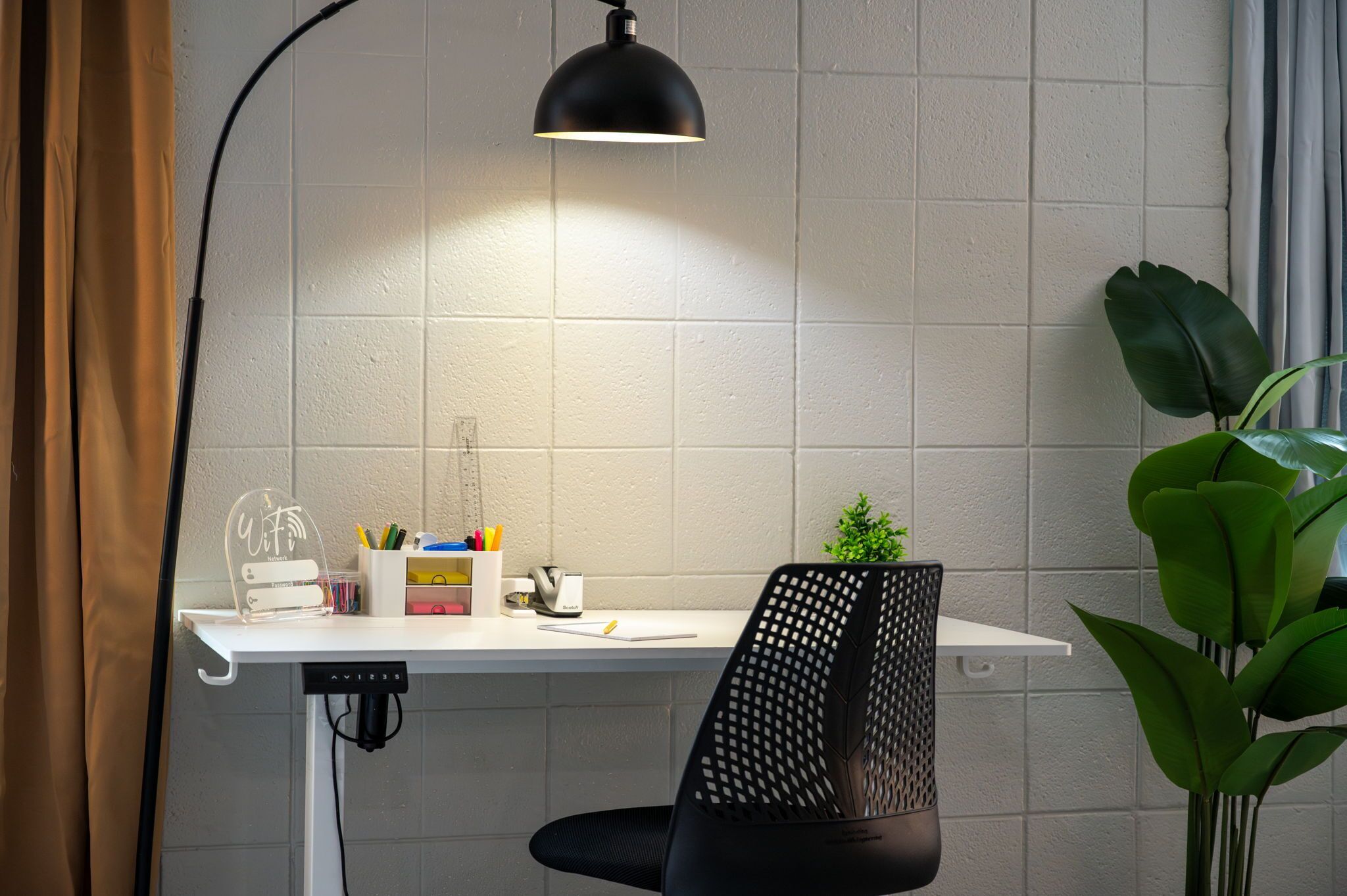 A modern workspace with a white desk, a black chair, a clear desk organizer, a potted plant, and a black pendant light.