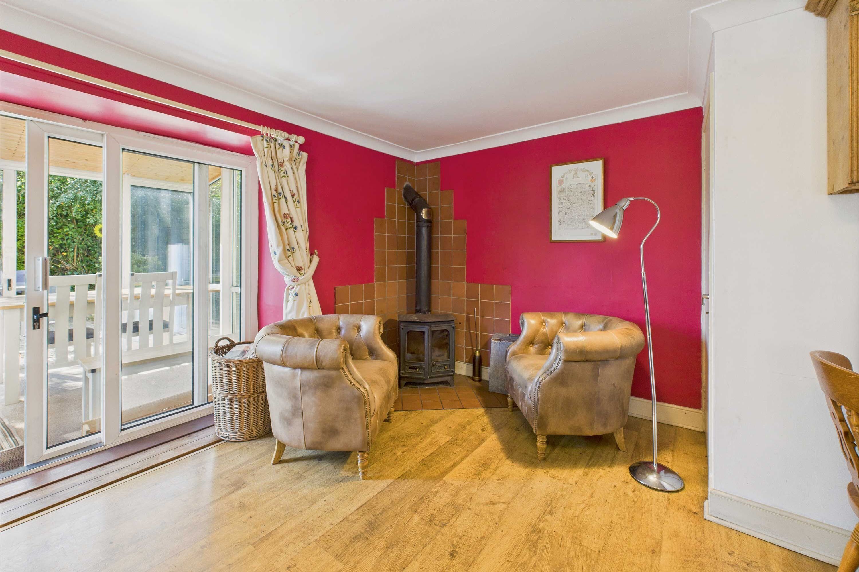 Una habitación con pared roja, dos sillones de cuero, una lámpara de pie, suelo de madera, una ventana con cortina y un pequeño calefactor.