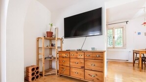 A room with a wooden dresser, a television mounted on the wall, a window with a view of greenery, and a small table with chairs. - Central, quiet oasis with garden and parking lot (Graz)