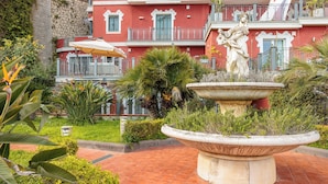 Un patio con una fuente, abundante vegetación y un edificio rojo con balcones.