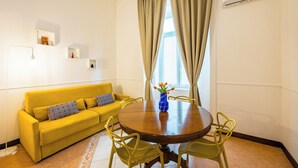 A room with a yellow sofa, a wooden dining table with chairs, a vase with flowers, and an air conditioning unit on the wall. - La Casa di Famiglia by Napoliapartments (Naples)