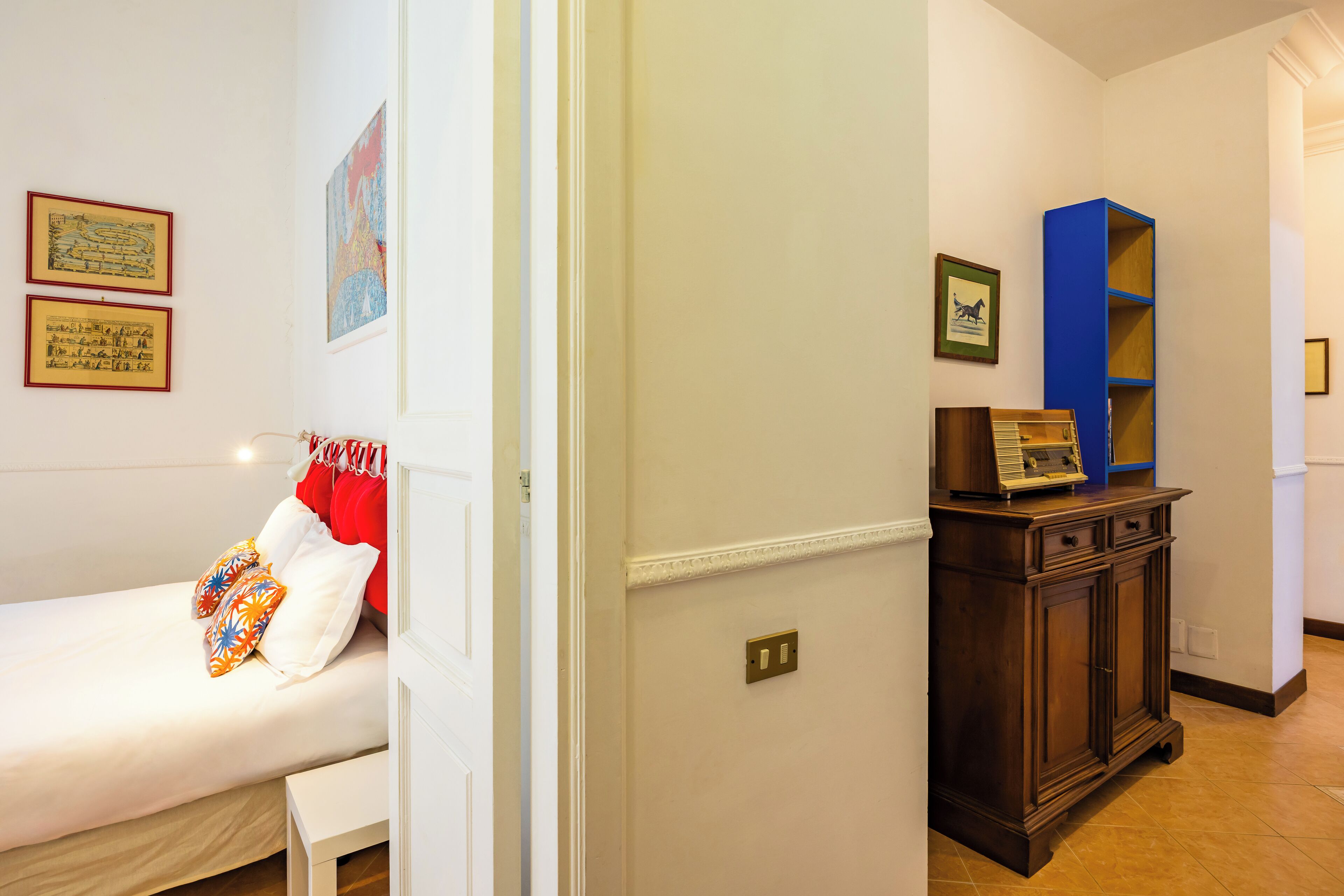 A bedroom with a bed, a bedside table, a nightstand, a wardrobe, and a shelf with books and decorative items.