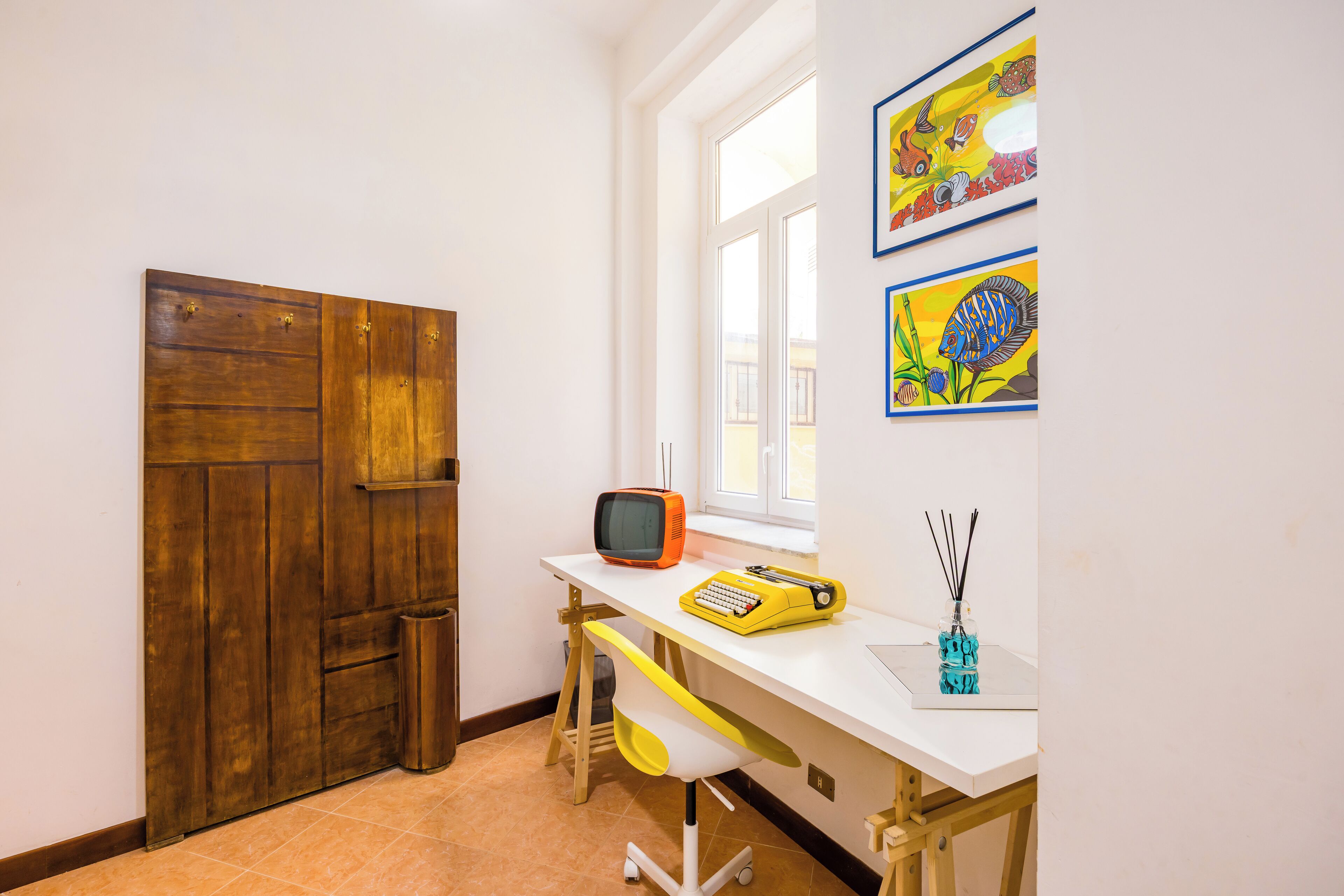Een kamer met een houten deur, een wit bureau met een geel typmachien, een kleine televisie en een raam met kunst aan de muur.
