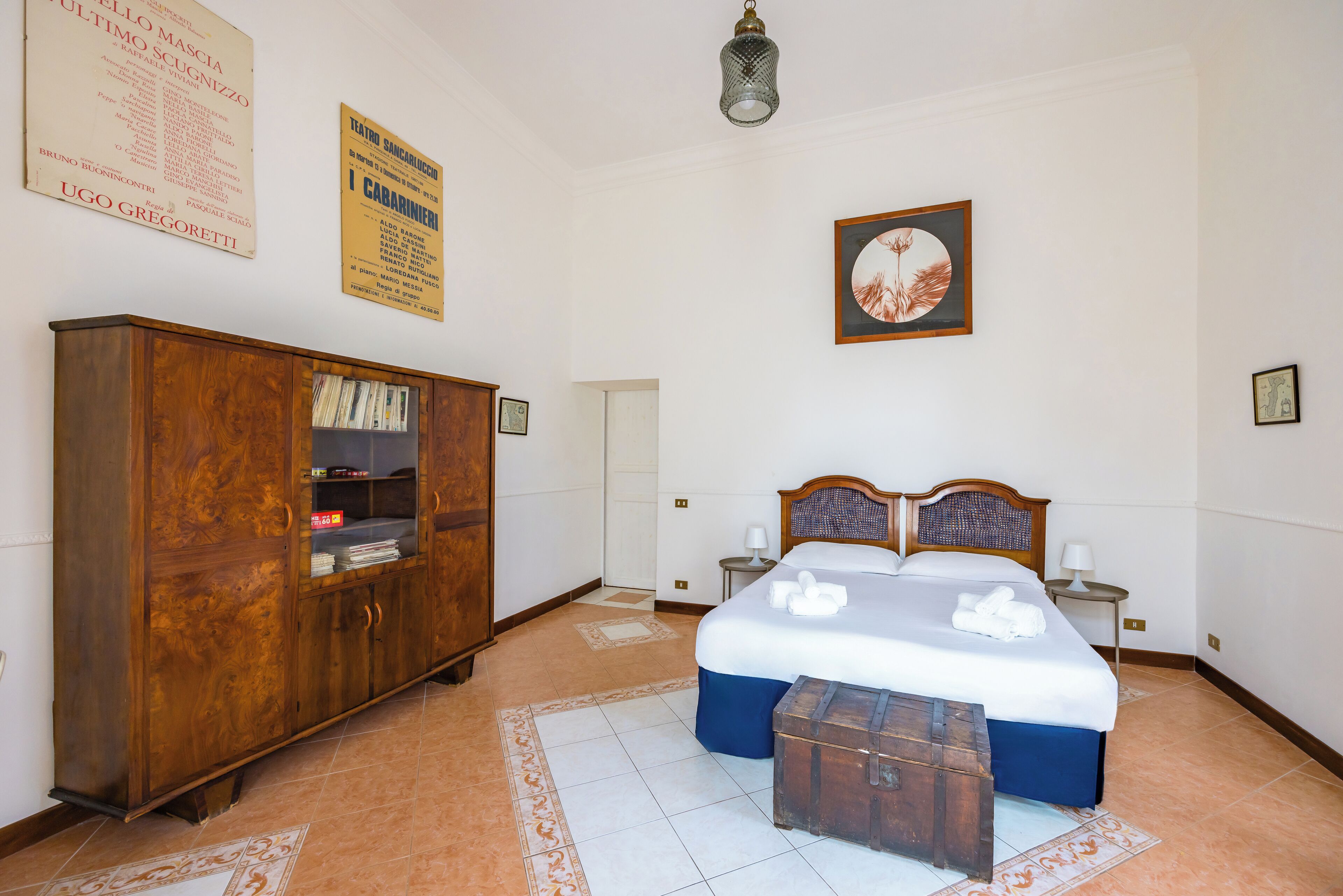 A hotel room with a large wooden wardrobe, a bed with a blue bedspread, a small wooden chest, and a framed picture on the wall.