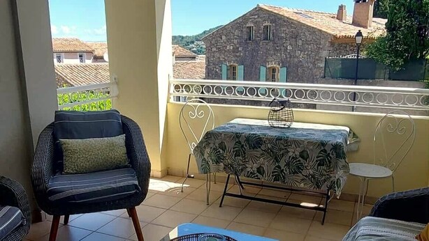 A balcony with a wicker table, two chairs, a hanging light fixture, and a view of a stone building.