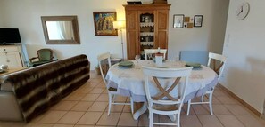 Un comedor con una mesa redonda dispuesta para cuatro personas, rodeada de sillas blancas. Un sofá marrón está ubicado contra la pared, y se observa un aparador con puertas de vidrio al fondo.