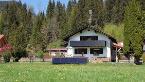 Ein zweistöckiges Haus mit Balkon, umgeben von einem Feld gelber Blumen und hohen Bäumen.