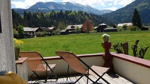 Ein Balkon mit zwei Korbstühlen und einem Topfpflanzen, mit Blick auf ein grünes Feld und Häuser, im Hintergrund Berge.