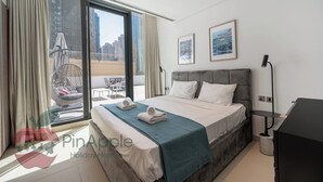 A modern hotel room with a large bed, a view of the city through floor-to-ceiling windows, a balcony with outdoor furniture, and a desk with a lamp. - Spacious 2 Bedroom With Maid Room Near Marina Walk (Dubai)