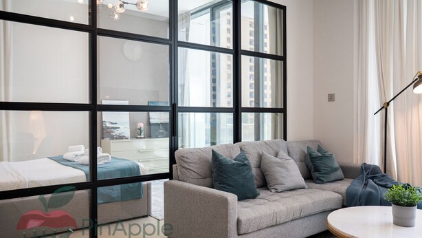 A modern living room with a grey sofa, a round coffee table, and a large window with a view of buildings. - Spacious 2 Bedroom With Maid Room Near Marina Walk (Dubai)