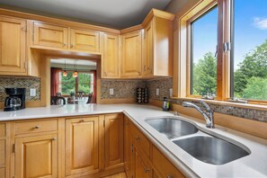 A kitchen with wooden cabinets, a double sink, a coffee maker, and a view of greenery outside.