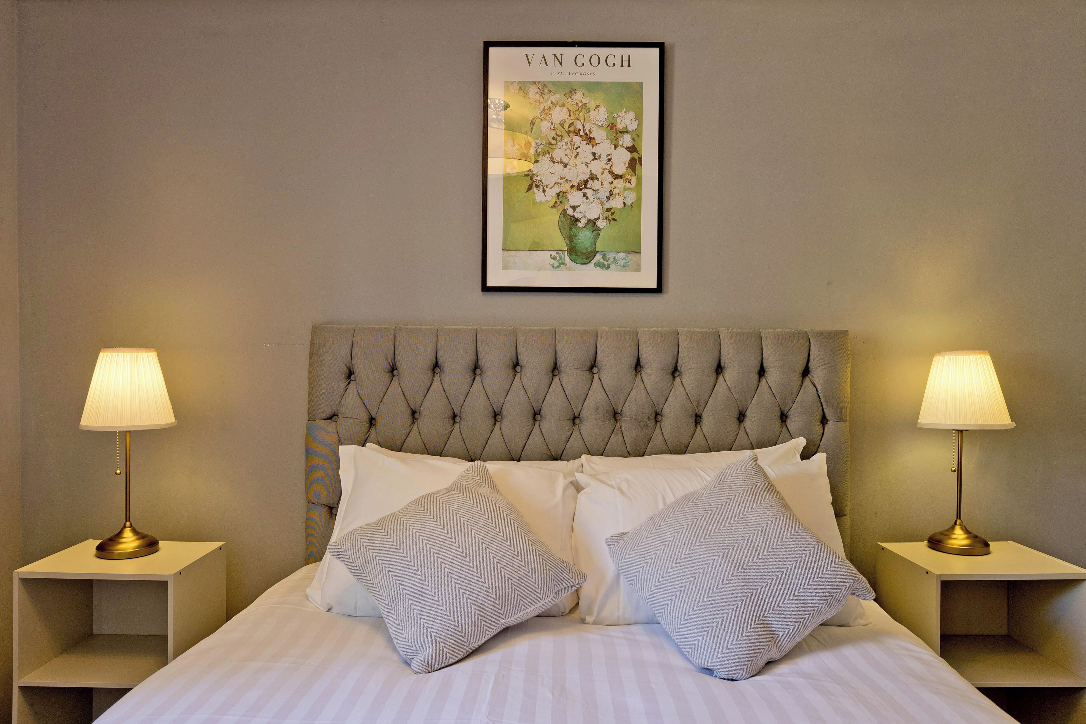 Een net opgemaakt bed met twee kussens, een ingelijst kunstwerk erboven en twee bedlampen op nachtkastjes.
