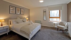 A modern bedroom with a bed, bedside tables, a chair, a small table, and a window with blinds. - Imperial Meridian Place Apartment (London)