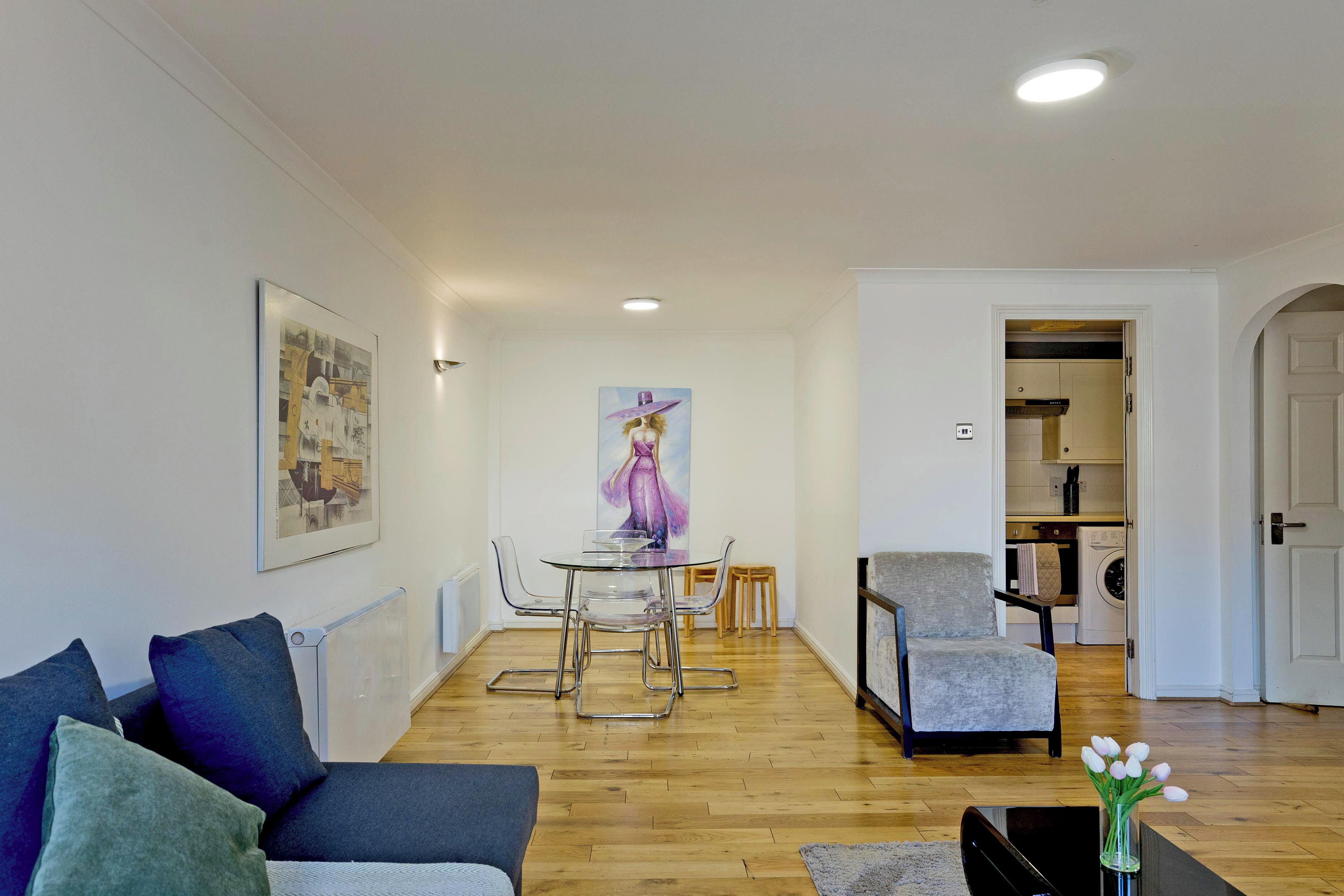 A modern living room with a sofa, a dining table with chairs, a painting on the wall, and a kitchen in the background.