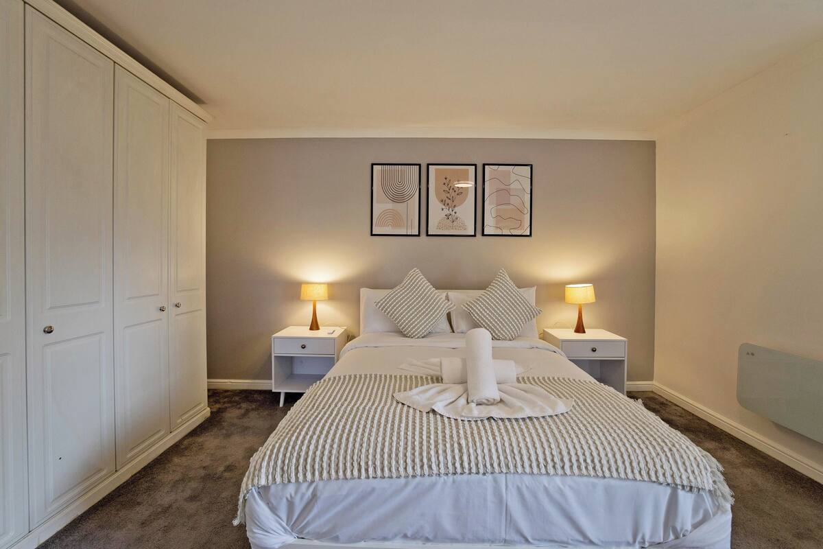 A neatly arranged bedroom with a large bed, two bedside tables with lamps, a wardrobe, and three framed artworks on the wall.