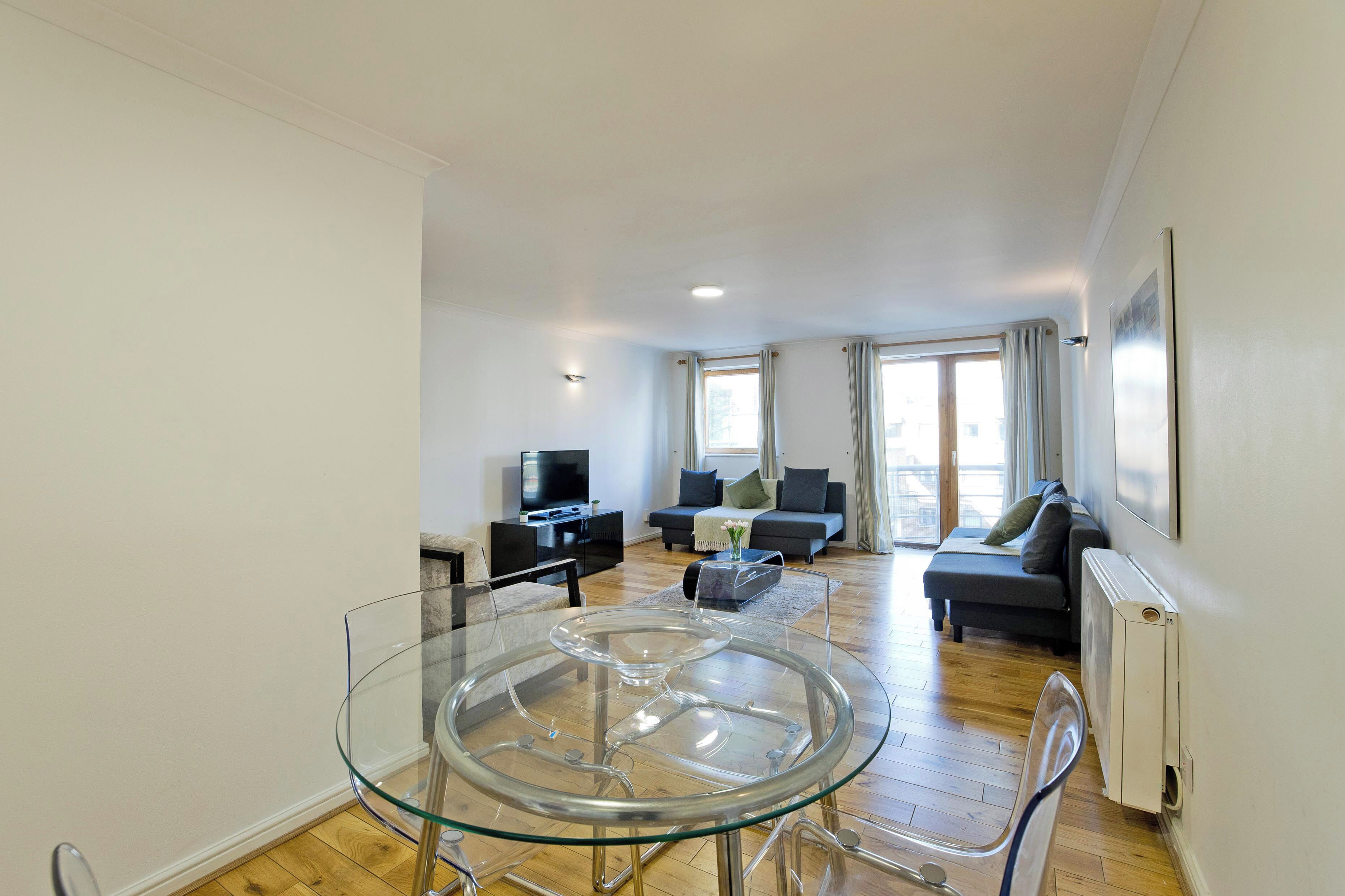 A modern living room with a glass dining table, a sofa set, a TV, and a radiator.