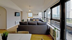 A modern living room with a grey sofa, a dining table, and large windows offering a city view. - Imperial Meridian Place Apartment (London)
