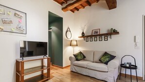 A living room with a TV on a wooden stand, a sofa with decorative pillows, a lamp, and a shelf with pictures and small plants. - Porta Romana Charme Flat - Florence Dolce Vita (Florence)