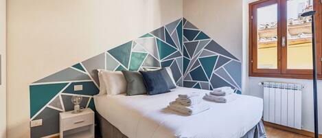 Une chambre avec un mur orné d’un motif géométrique, un lit recouvert de draps blancs, une table de chevet et une fenêtre donnant sur des immeubles.