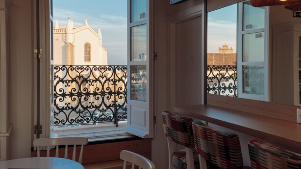 An open window with a view of a building, a balcony with a wrought iron railing, and a table with chairs. - Heaven Inn Suites & Terrace (Évora)