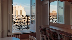 An open window with a view of a building, a balcony with a wrought iron railing, and a table with chairs.