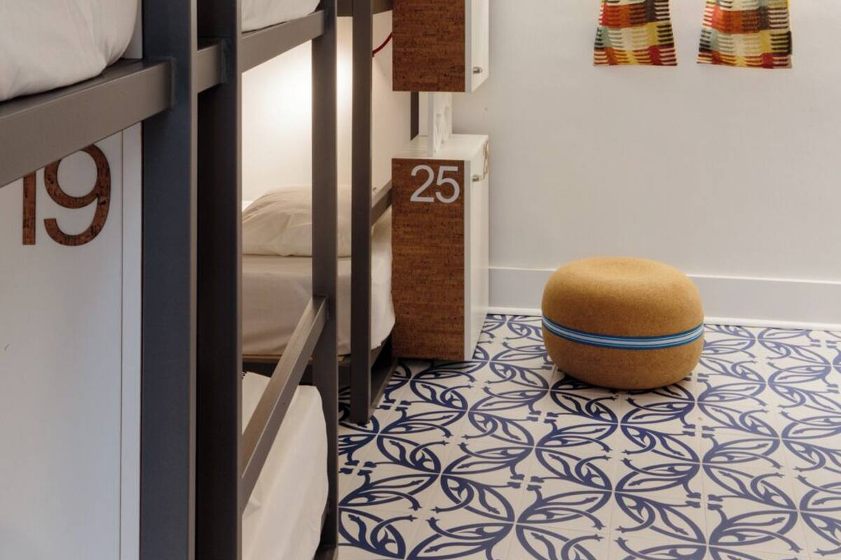 Une chambre d’hôtel avec des lits superposés, des compartiments de rangement numérotés, un sol à motifs et un petit pouf.