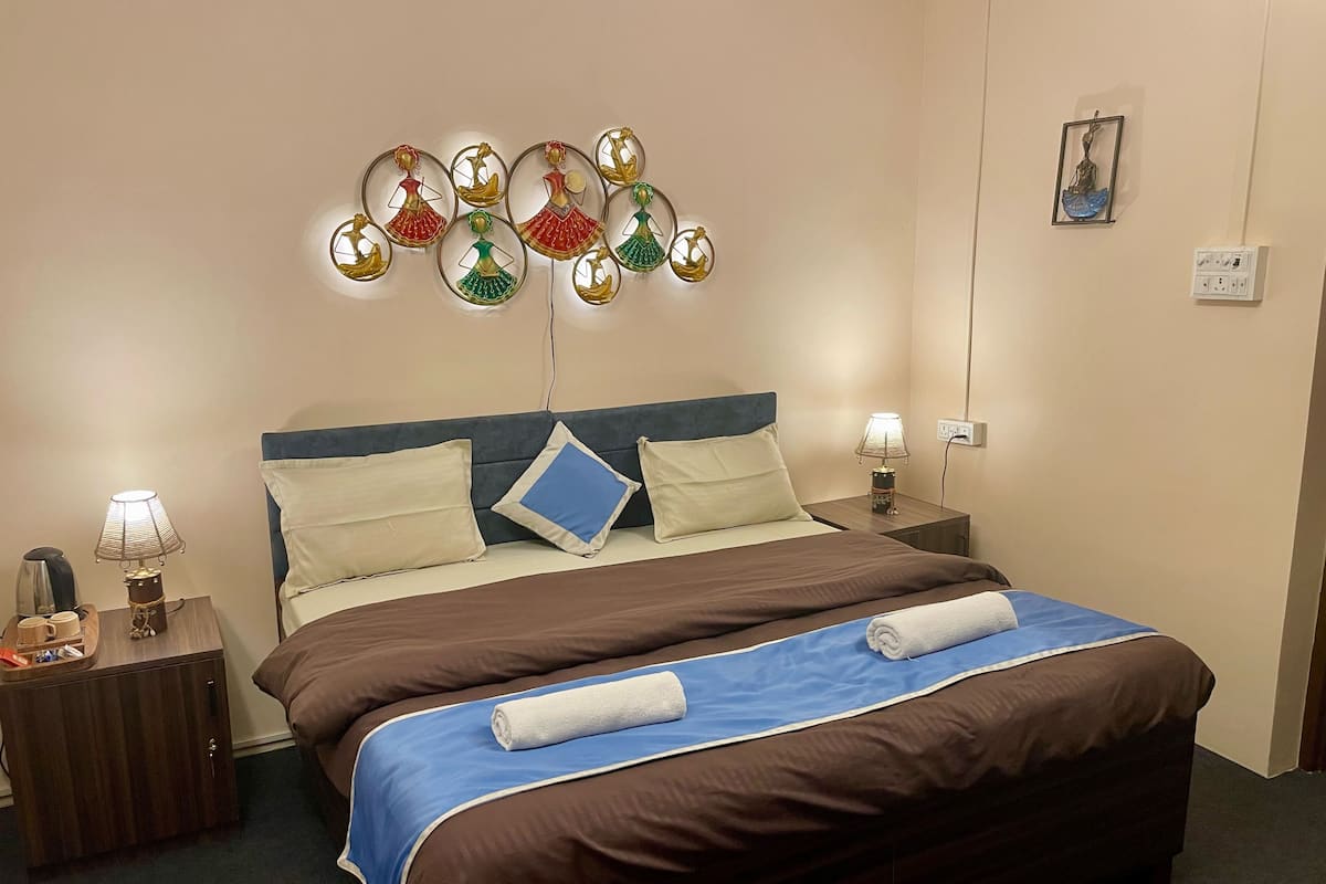 Un letto ben rifatto con copripiumino blu e marrone, due cuscini e un asciugamano arrotolato. Sono presenti due comodini con lampade e un piccolo oggetto decorativo.
