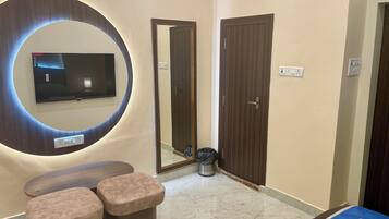 Habitación de hotel con un espejo grande, un televisor de pantalla plana, una cama con cubrecama azul y un pequeño espacio de descanso con dos taburetes acolchados.