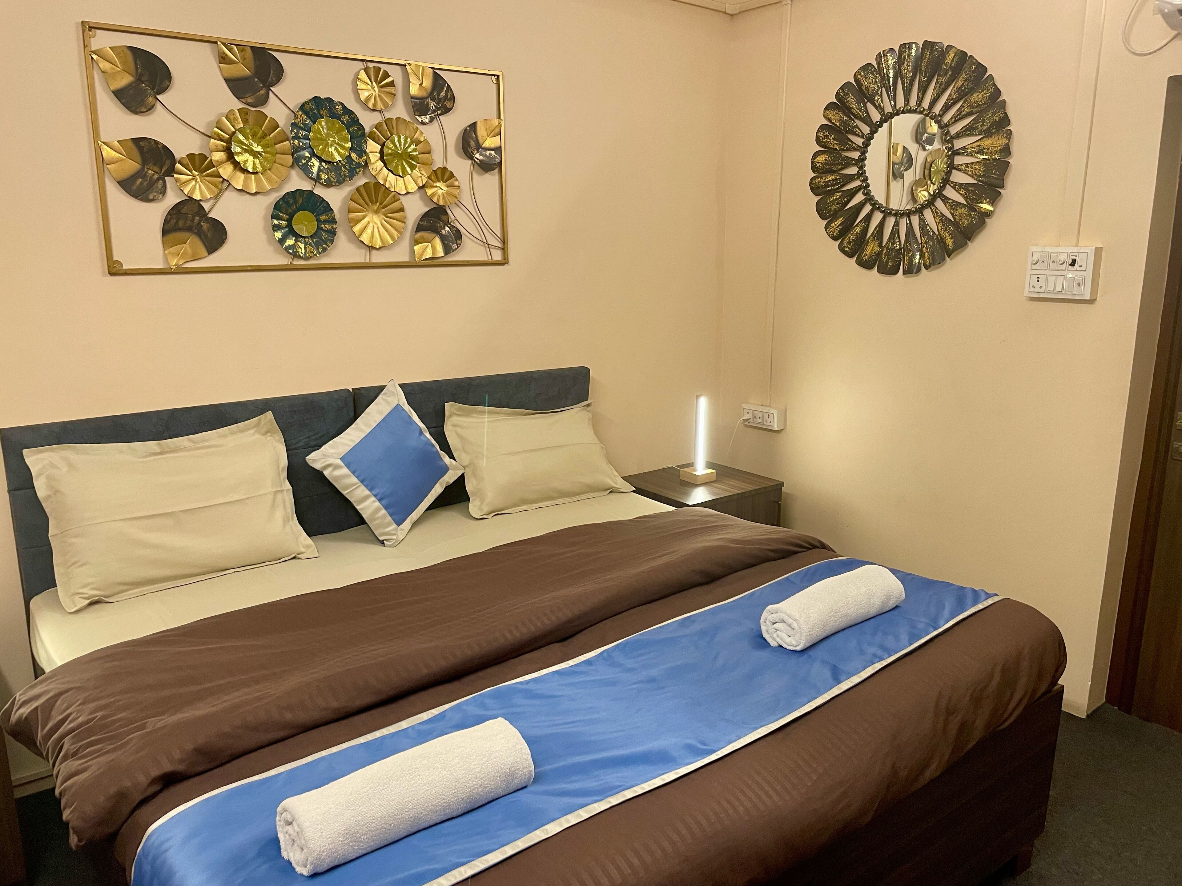 A neatly made bed with a blue and brown bedspread, two pillows, and two rolled towels. A decorative wall art piece and a wall clock are visible.