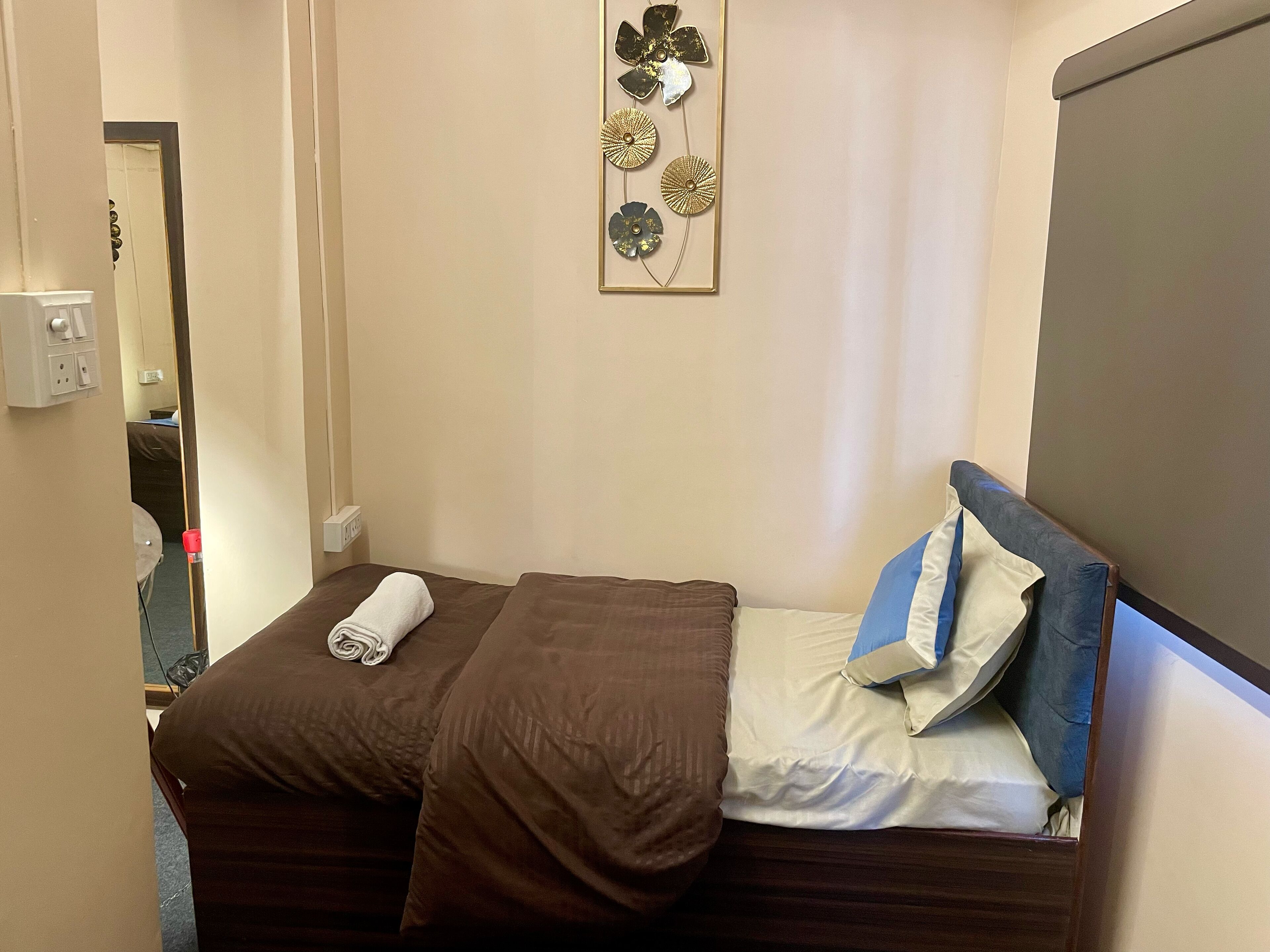 Uma cama pequena e bem arrumada com roupa de cama marrom e uma toalha enrolada, um travesseiro azul e branco, e um objeto decorativo na parede acima.