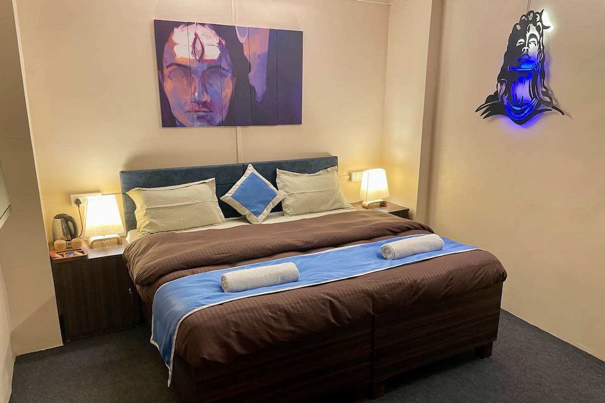 Un letto rifatto con precisione, con una testata marrone, una coperta blu e due asciugamani bianchi. Si notano due lampade da comodino, un comodino con una lampada e un'opera d'arte appesa al muro.