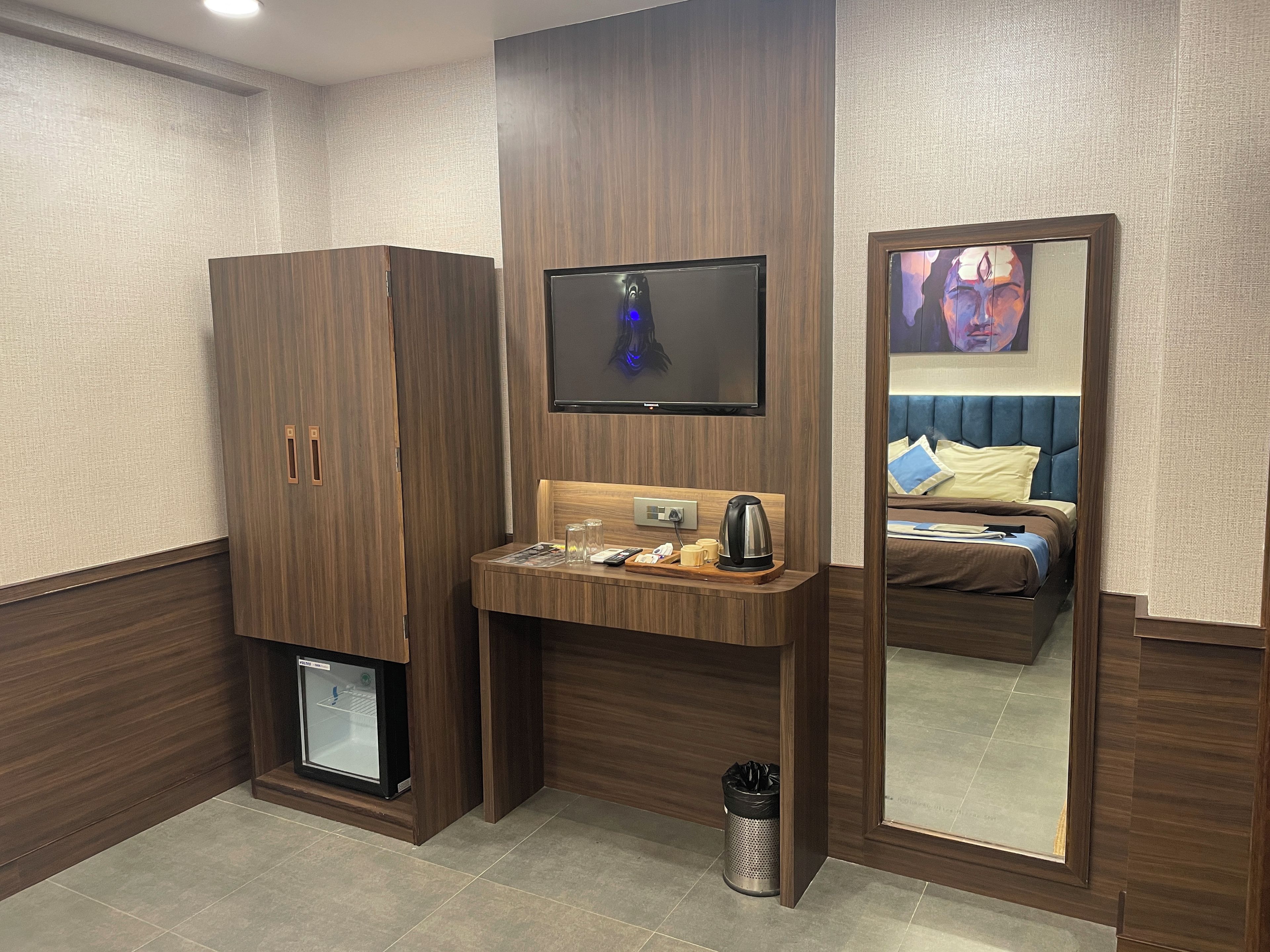 A hotel room with a wooden wardrobe, a small sink with a kettle, a trash bin, and a bed with blue headboard and pillows.