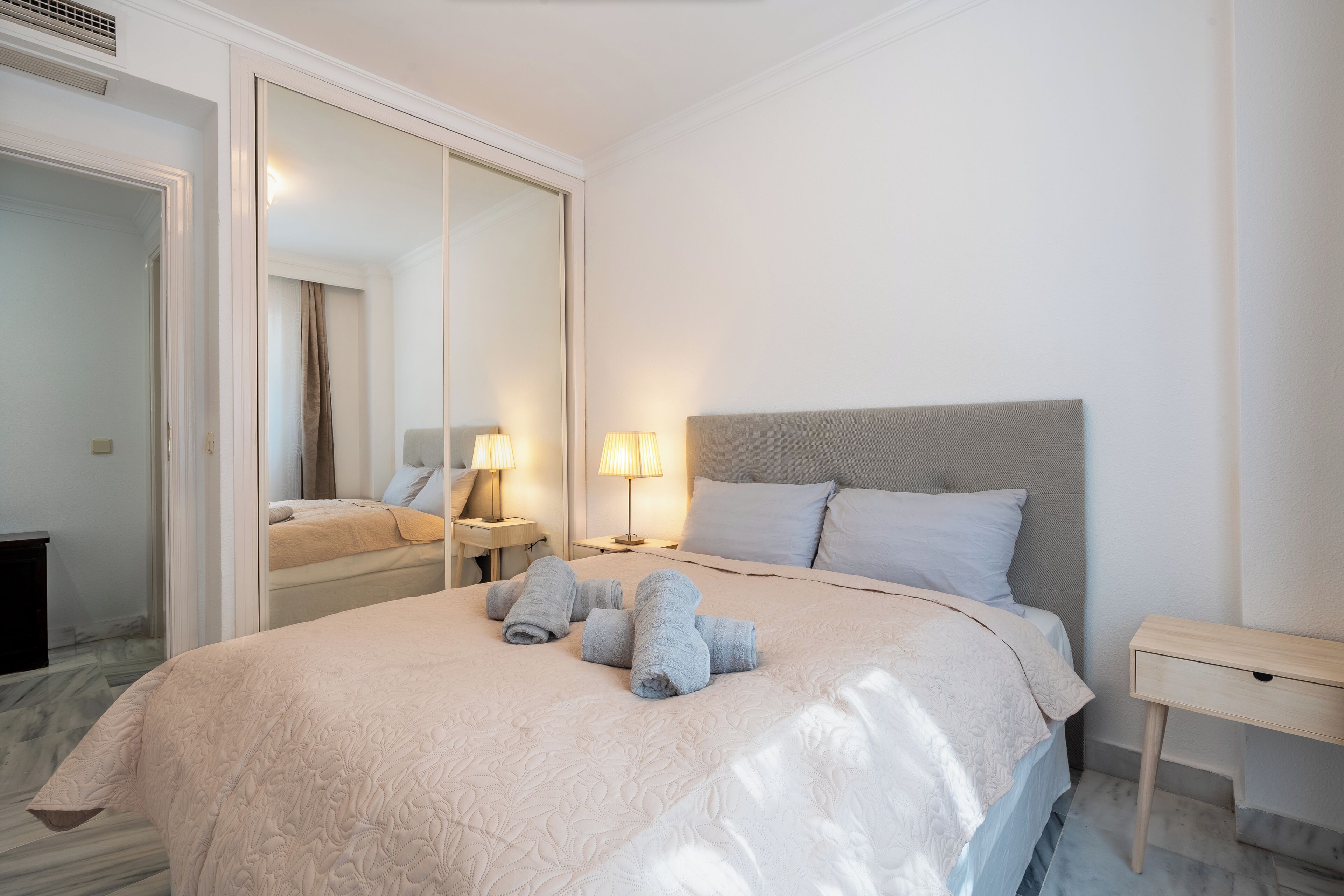 A neatly made bed with a light-colored comforter and pillows, a bedside table with a lamp, and a large mirror on the wall.