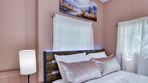 Un dormitorio con cabecera de madera, ropa de cama blanca, una lámpara de noche y un cuadro sobre la ventana.