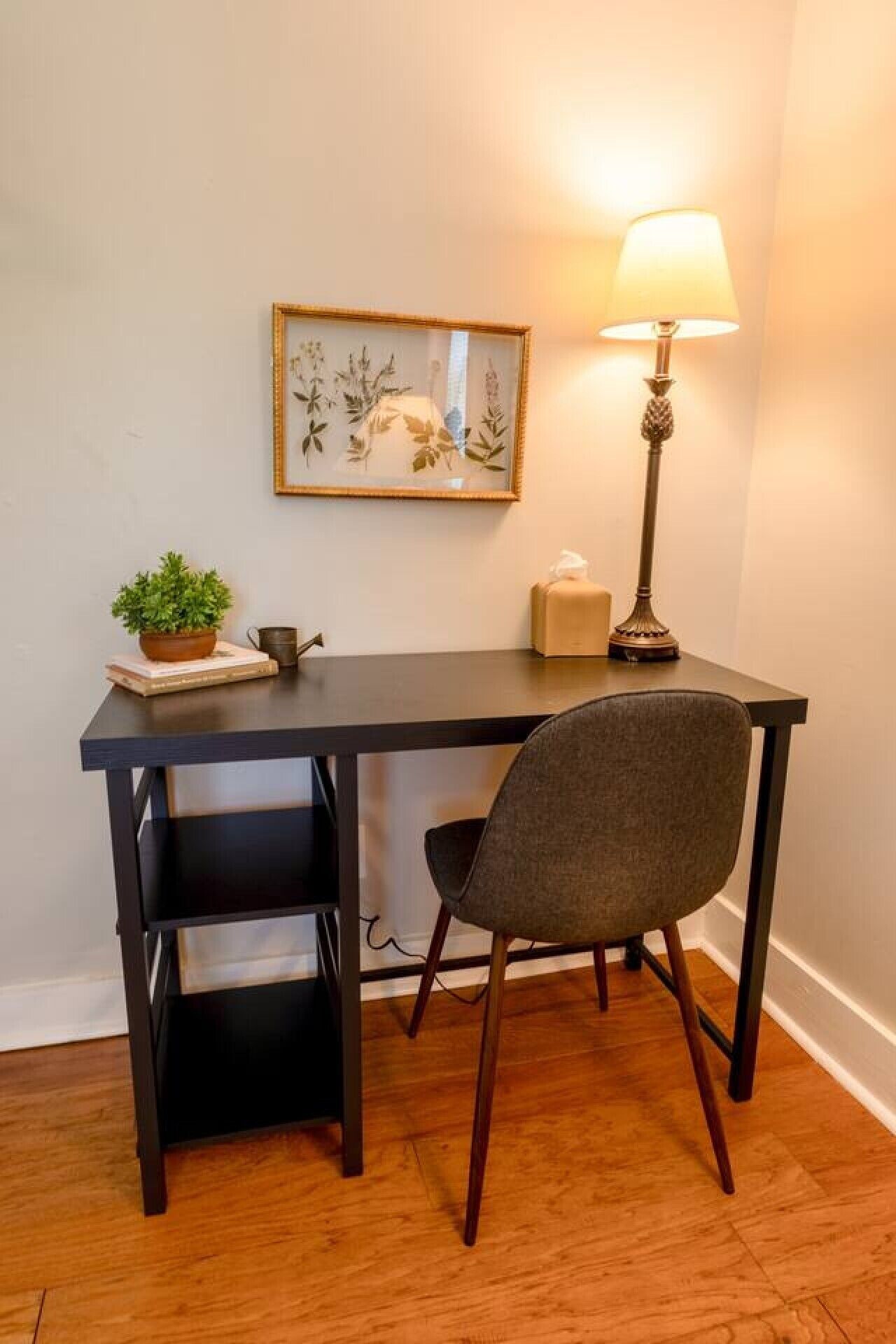 A dark wooden desk with a chair, a framed botanical print, a lamp, and a small plant on the desk.