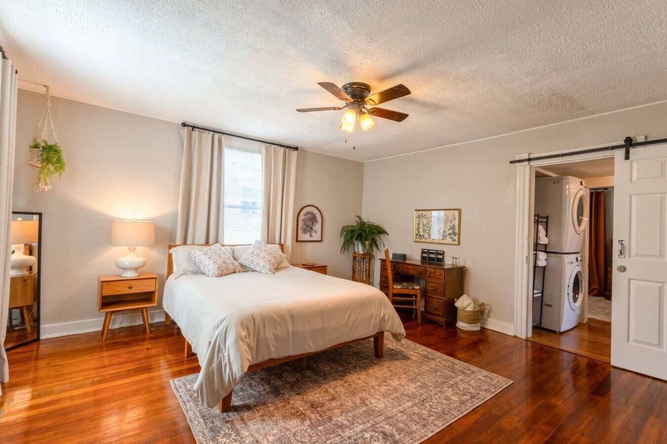 A bedroom with a bed, a ceiling fan, a desk, a chair, a lamp, a hanging plant, and a washing machine.