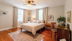 Ein Schlafzimmer mit Bett, Deckenventilator, Schreibtisch, Stuhl, Schreibtischlampe, Pflanze und einem gerahmten Bild an der Wand.