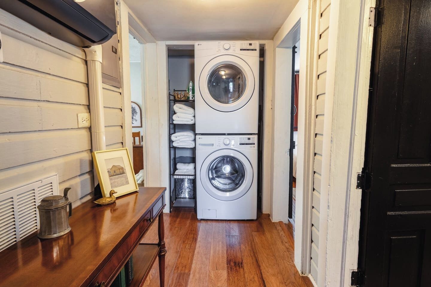 A laundry room with two stacked front-loading washing machines, a wooden table with a framed picture, and a shelf with folded towels.