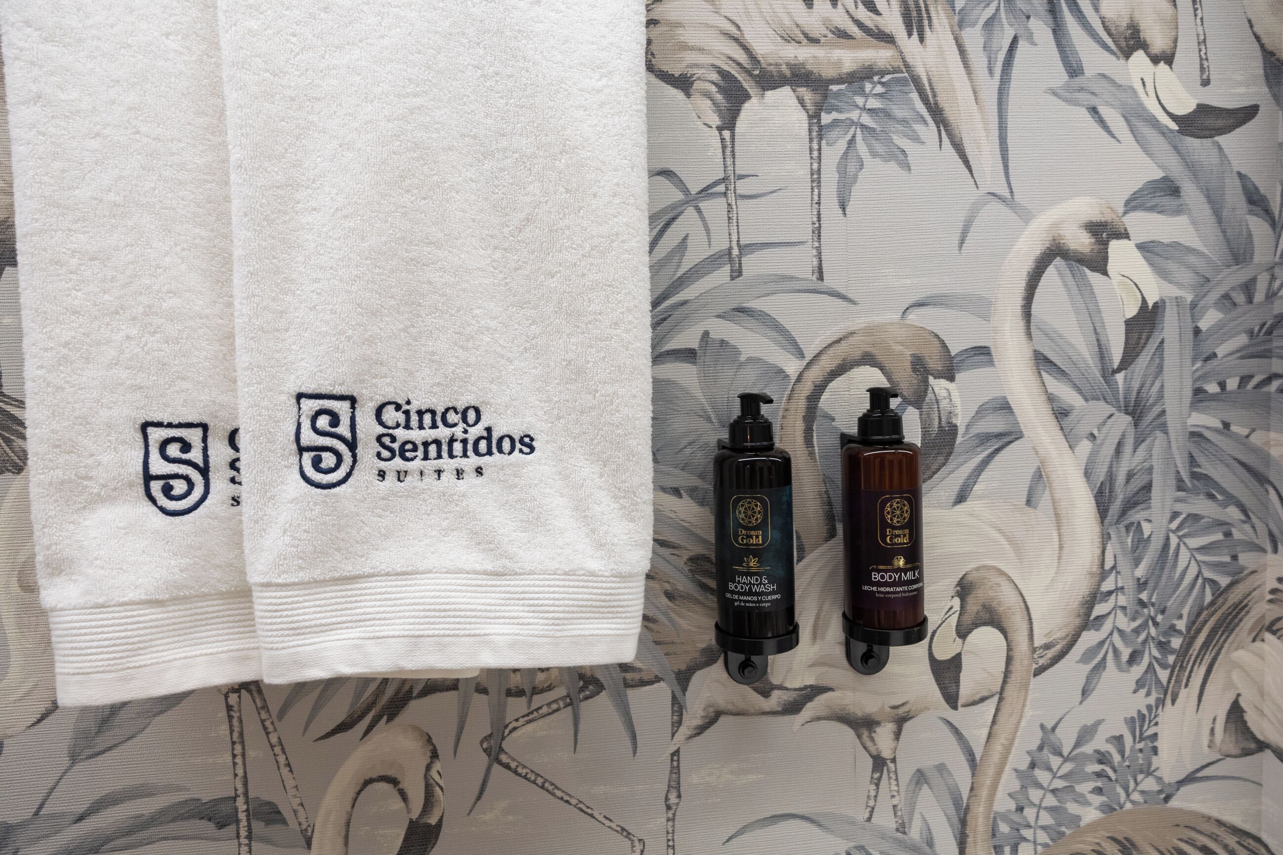 Toalla blanca con el logotipo de 'Cinco Sentidos Suites' y dos productos de cuidado corporal sobre un revestimiento de pared estampado.