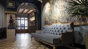A room with a tufted grey sofa, a decorative wall with a floral pattern, a large plant, and a clock on the wall. - 5 Sentidos - Suites de Lujo (Tudela)